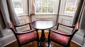 Une table ronde en bois, accompagnée de deux chaises rembourrées, est placée devant une fenêtre à cadre blanc, offrant une vue sur la verdure à l’extérieur.