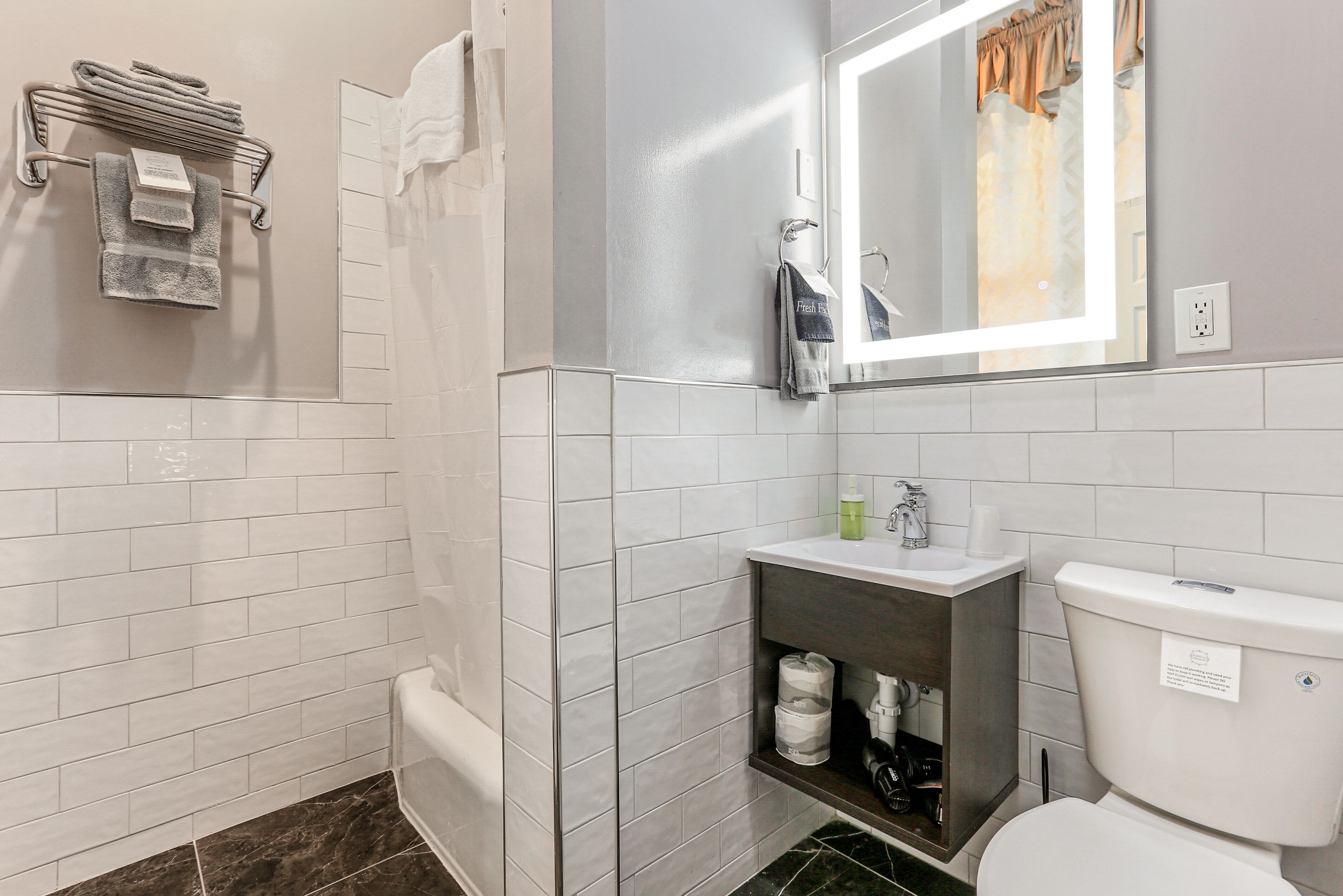 Un baño con paredes de azulejos blancos, una bañera blanca, un lavamanos con espejo y un inodoro.