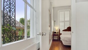A narrow hallway leading to a bedroom with a white bed, a wooden floor, and a window with a metal gate.