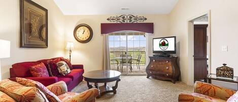 A living room with a red sofa, patterned armchairs, a coffee table, a clock, a television, and a balcony with a view.