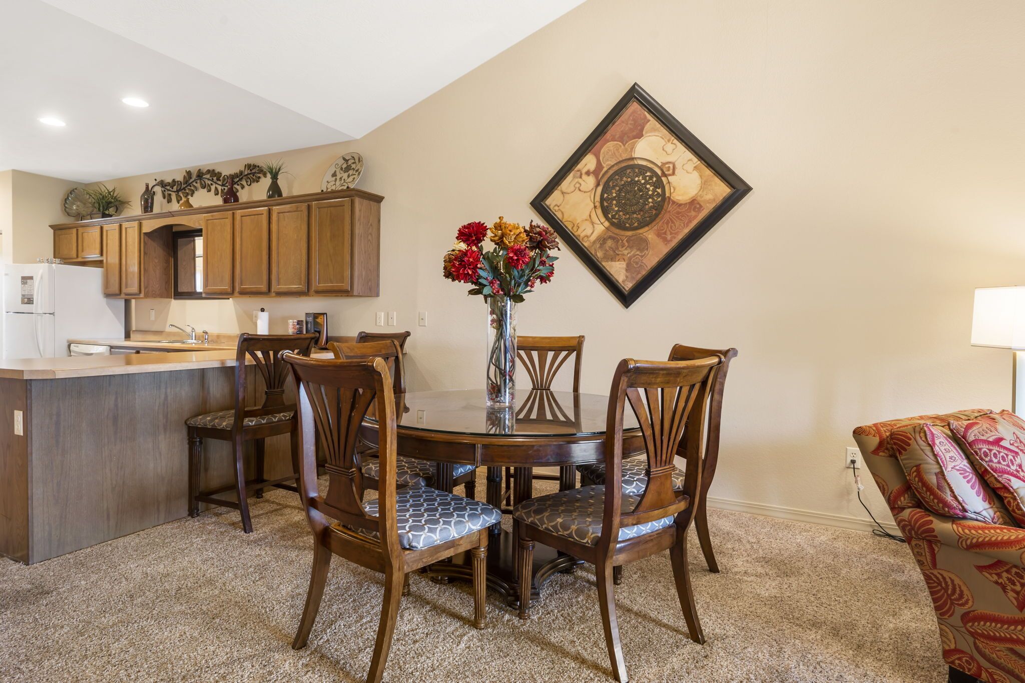 A dining area with a round table, four chairs, a vase with flowers, wooden cabinets, and a kitchen in the background.