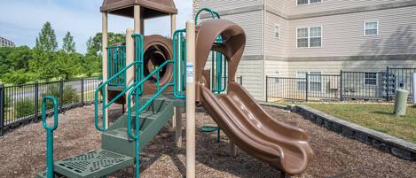 A playground with a slide and climbing structures, surrounded by a fence and a building in the background.