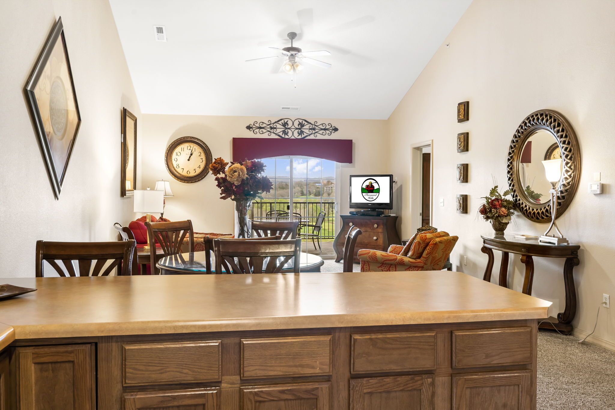 A dining area with a large wooden table, chairs, a ceiling fan, and a view of the outdoors through a window with a red curtain.