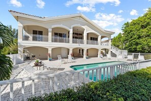 Une maison à deux étages avec une piscine, un espace extérieur aménagé pour s’asseoir et une clôture blanche.