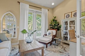 Una habitación con una mesa de madera, un sillón blanco, un sofá y un ventanal con cortinas blancas.