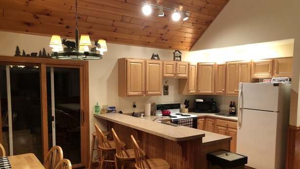 A kitchen with wooden cabinets, a central island with bar stools, a refrigerator, and a stove. - Hideaway in the Great North Woods - direct access to snowmobile trails (Errol)