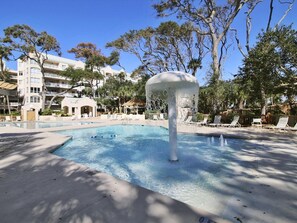 A swimming pool with a water fountain, lounge chairs, and a building with balconies in the background.
