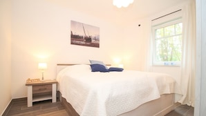 A neatly arranged bedroom with a bed, bedside lamps, a nightstand, a window with curtains, and a wall-mounted picture.