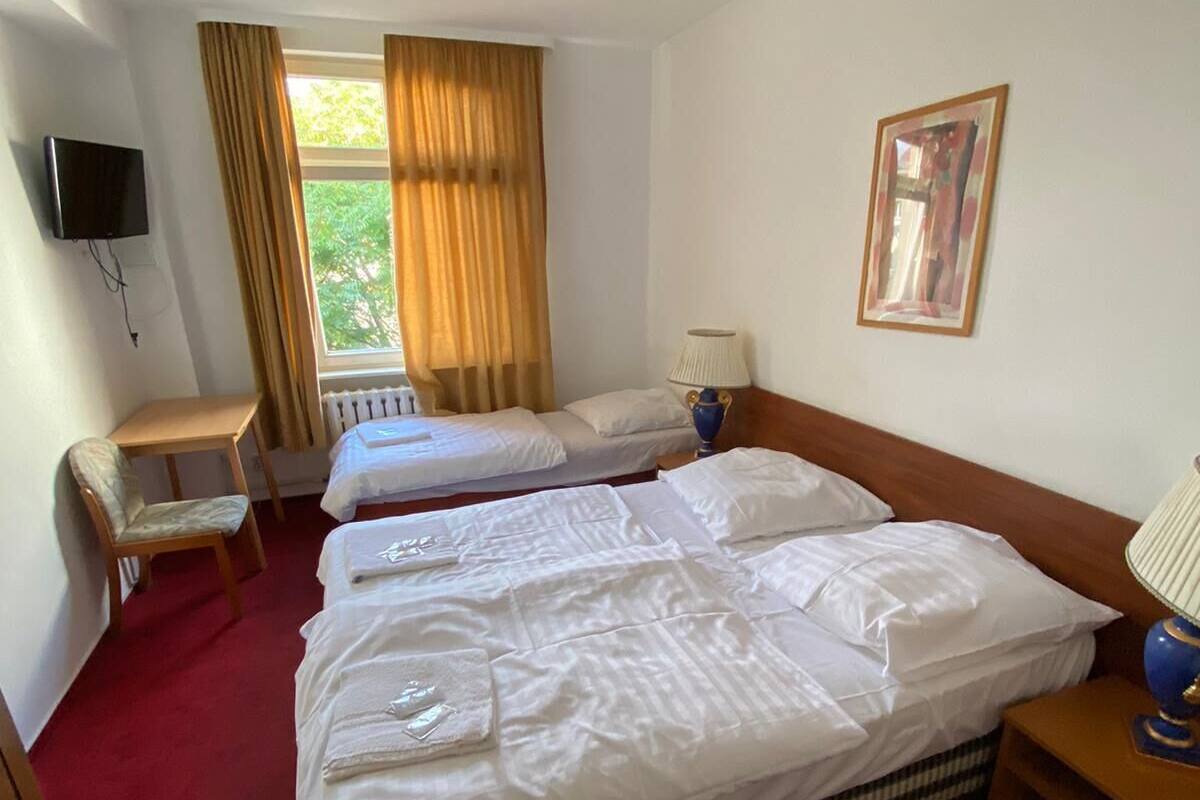 A hotel room with two single beds, a small table, a chair, a TV mounted on the wall, and a window with curtains.