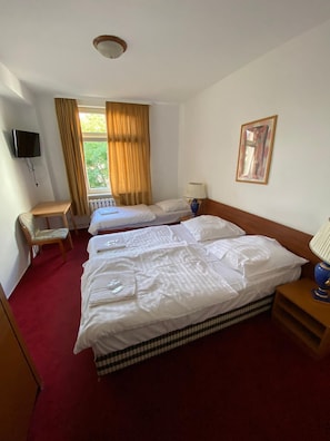 Habitación de hotel con dos camas individuales, una mesita pequeña, una silla, un televisor montado en la pared y una ventana con cortinas.