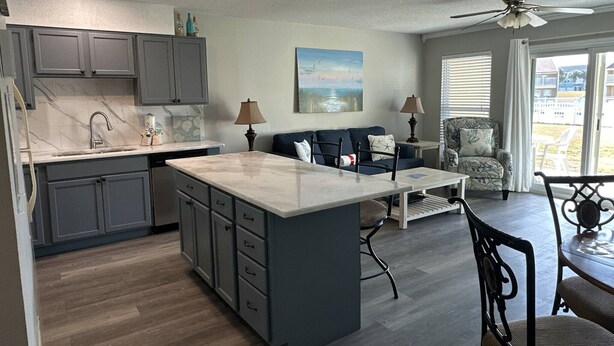 A modern kitchen with grey cabinets, a marble countertop, and a central island. There is a dining area with a table and chairs, a sofa, and a chair with a patterned cushion. A ceiling fan and a window with blinds are visible. - 9A, Resting Beach Face (Destin)
