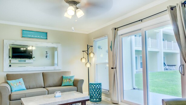 A living room with a sofa, coffee table, and a TV mounted on the wall. There is a sliding glass door leading to a balcony with a view of a house and a lawn. - 7A, Sun Sand & Solitude (Destin)