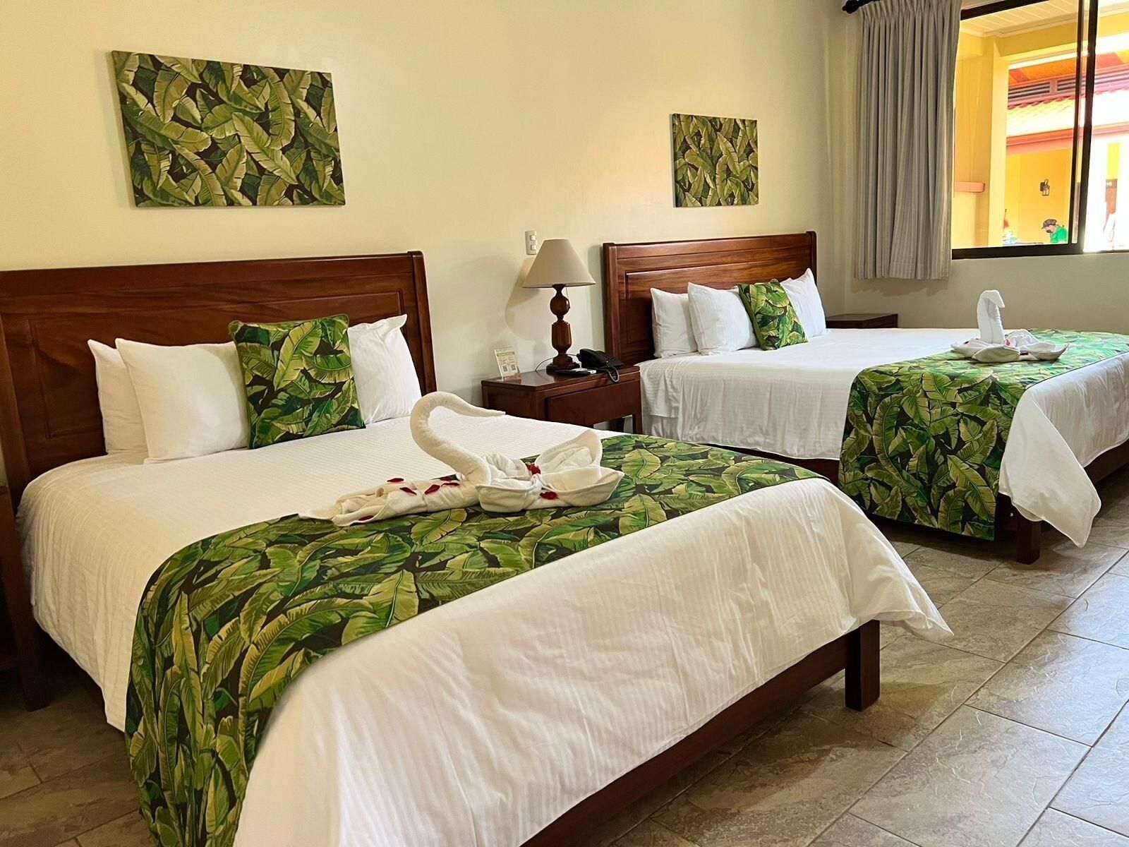 A hotel room with two beds, each with a green leaf-patterned bedspread, white linens, and a towel on the bed. There are two paintings on the wall, a lamp on the nightstand, and a window with curtains.