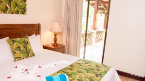 A hotel room with a bed, a nightstand, a lamp, a balcony with a railing, and a view of the outside. - Rancho Casa Grande (Quepos)