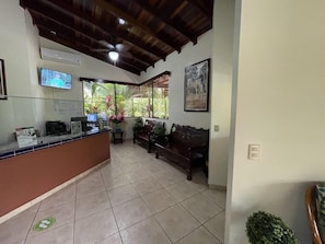 A reception area with a wooden ceiling, a reception desk, a sofa, and a window with a view of greenery. - Rancho Casa Grande (Quepos)