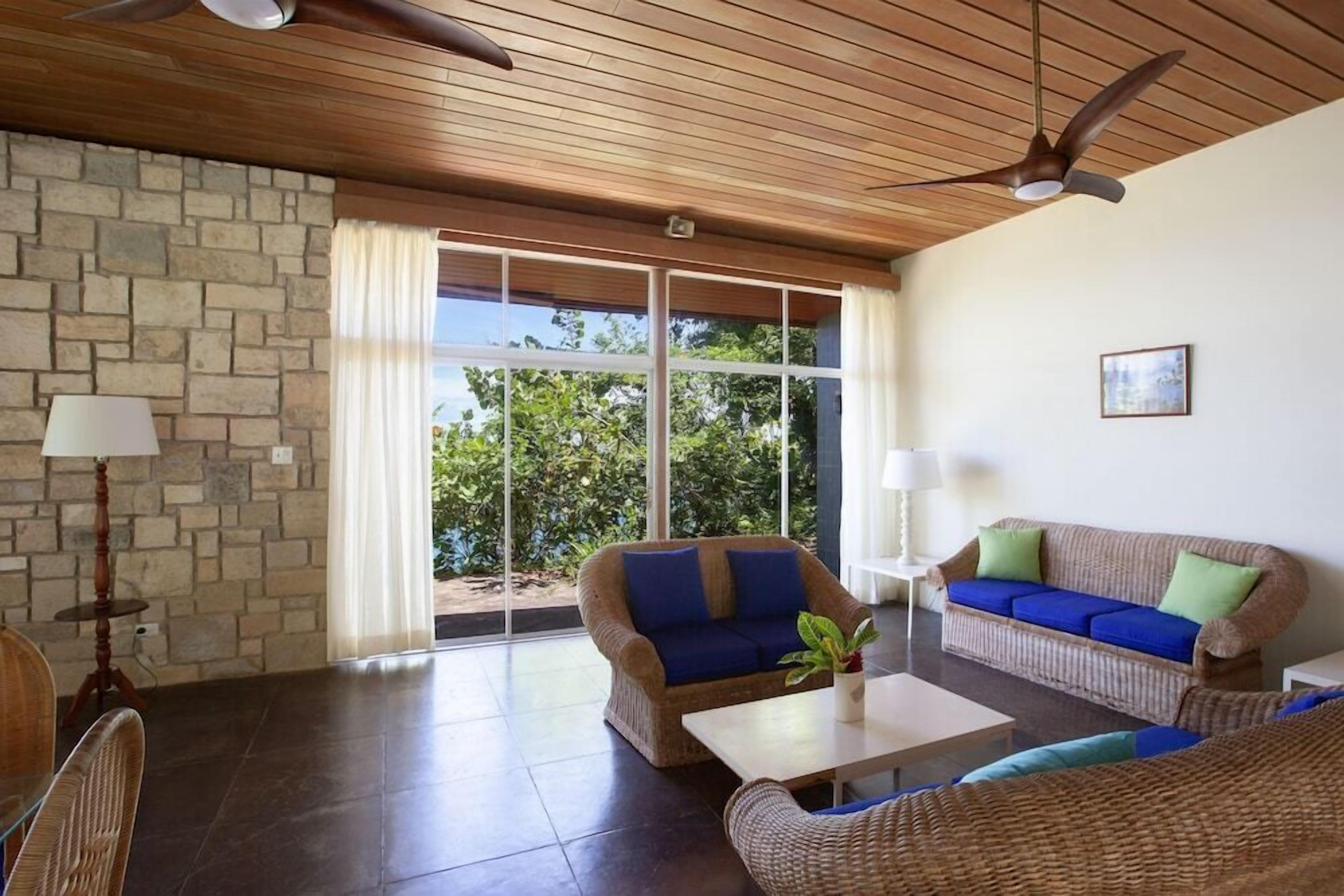 A spacious living area with wicker furniture, a stone wall, and large windows offering a view of greenery.
