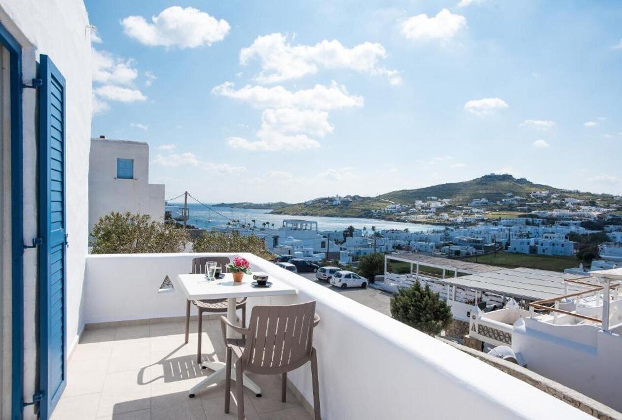 Un balcon avec une table et des chaises, donnant sur une ville côtière aux bâtiments blancs et offrant une vue sur la mer.