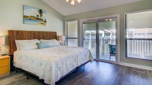 A bedroom with a bed, a ceiling fan, a painting, and a view of the outside.