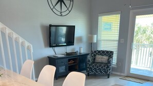A modern living room with a white marble table, white chairs, a black sofa with a patterned cushion, a television on the wall, and a large window with blinds. - 8F, Isle Be Back (Destin)