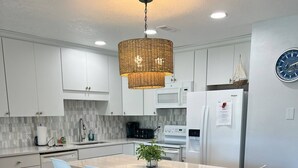 A modern kitchen with white cabinets, a central island with chairs, a hanging light fixture, and a clock on the wall. - 8F, Isle Be Back (Destin)