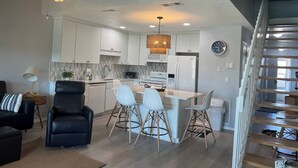 A modern kitchen with white cabinets, a central island with bar stools, a dining area with a black leather chair, and a staircase with wooden steps. - 8F, Isle Be Back (Destin)