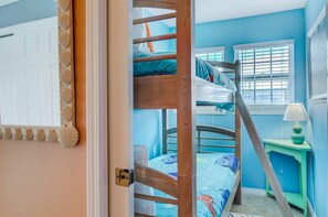 A room with a bunk bed, a small green nightstand with a lamp, and a window with blinds.