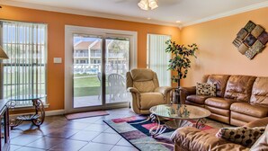 A living room with a brown leather couch, a glass coffee table, and a beige armchair. There is a large window with blinds, a ceiling fan, and a decorative wall hanging. - 4A, Castaway Cove (Destin)