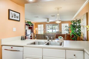 A kitchen with a white countertop, double sink, and built-in dishwasher. There is a dining area with wicker furniture and a ceiling fan.