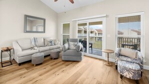 A spacious living room with a grey sofa, ottoman, and patterned armchair, a ceiling fan, and a large sliding glass door leading to a balcony. - 7E, Shell We Stay? (Destin)