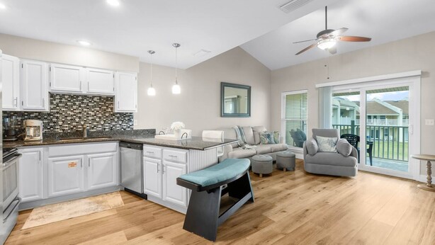 A modern kitchen with white cabinets, a black countertop, and a wooden floor. There is a dining area with a gray sofa and a ceiling fan. - 7E, Shell We Stay? (Destin)