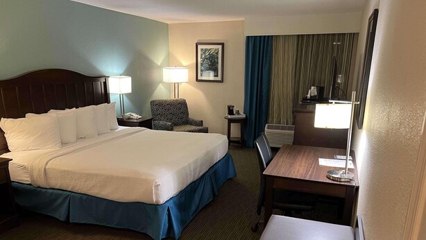 A hotel room with a large bed, a desk, a chair, a lamp, a small table, and a picture on the wall.