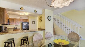 A kitchen with wooden cabinets, a refrigerator, and a dining area with a glass table and wicker chairs. - 3A, Rumrunner Retreat (Destin)