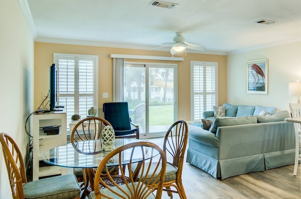 A living room with a glass-top dining table, wooden chairs, a blue armchair, a grey sofa, and a ceiling fan.