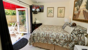 A bedroom with a bed, a desk, a chair, a TV, and a door leading to a balcony. - Fantasy Island Inn - Poolside Queen Room 3 (Wilton Manors)