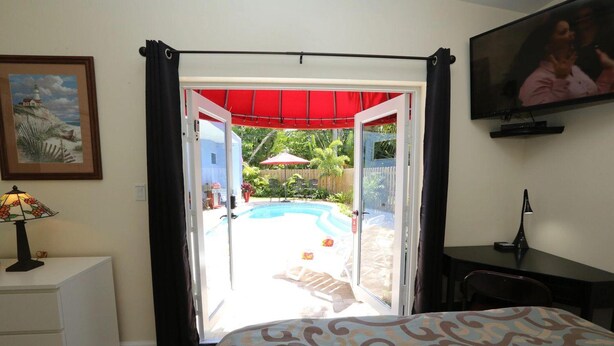 A bedroom with a bed, a desk, a TV, a lamp, and a view of an outdoor swimming pool. - Fantasy Island Inn - Poolside Queen Room 3 (Wilton Manors)