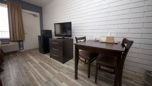 Habitació amb televisió, un petit armari, una taula i cadires, i una paret de maó blanc.