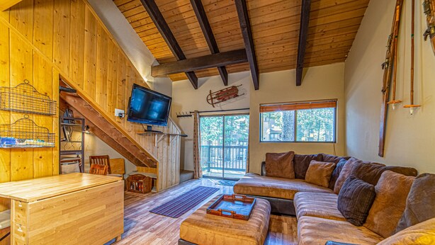 Interior acogedor de una cabaña con una mesa de comedor de madera, un sofá, un televisor y una escalera que lleva a un balcón.