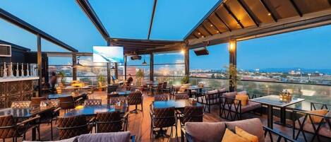 An outdoor seating area with tables and chairs, a bar, and a large TV screen.