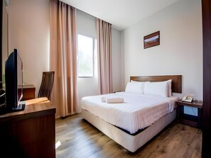 Ett hotellrum med en säng, ett skrivbord, en stol, en tv, ett fönster och en tavla på väggen.