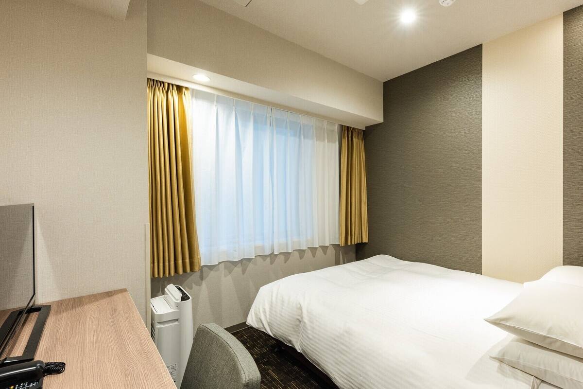 A hotel room with a bed, a desk, a chair, a telephone, and a window with curtains.