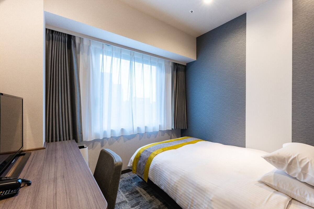A hotel room with a bed, a desk, a chair, a TV, and a window with curtains.
