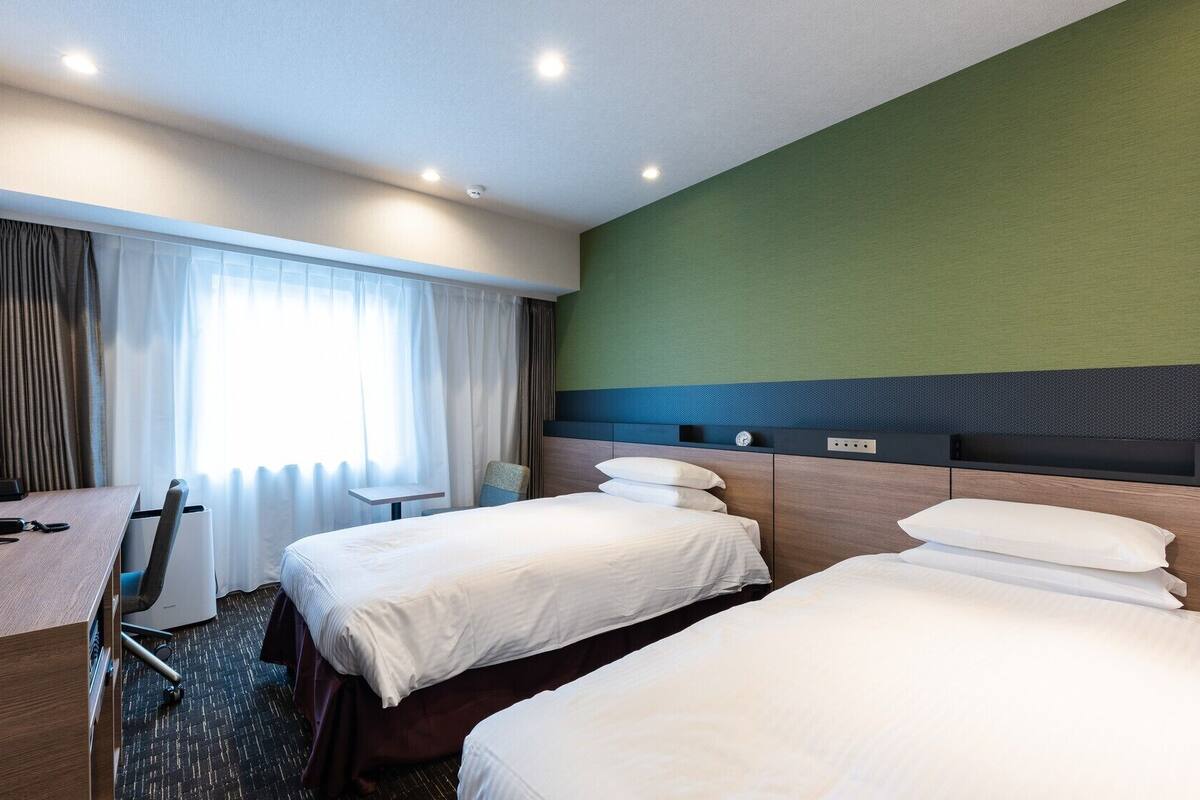 A hotel room with two beds, a desk, a chair, a TV, and a window with curtains.