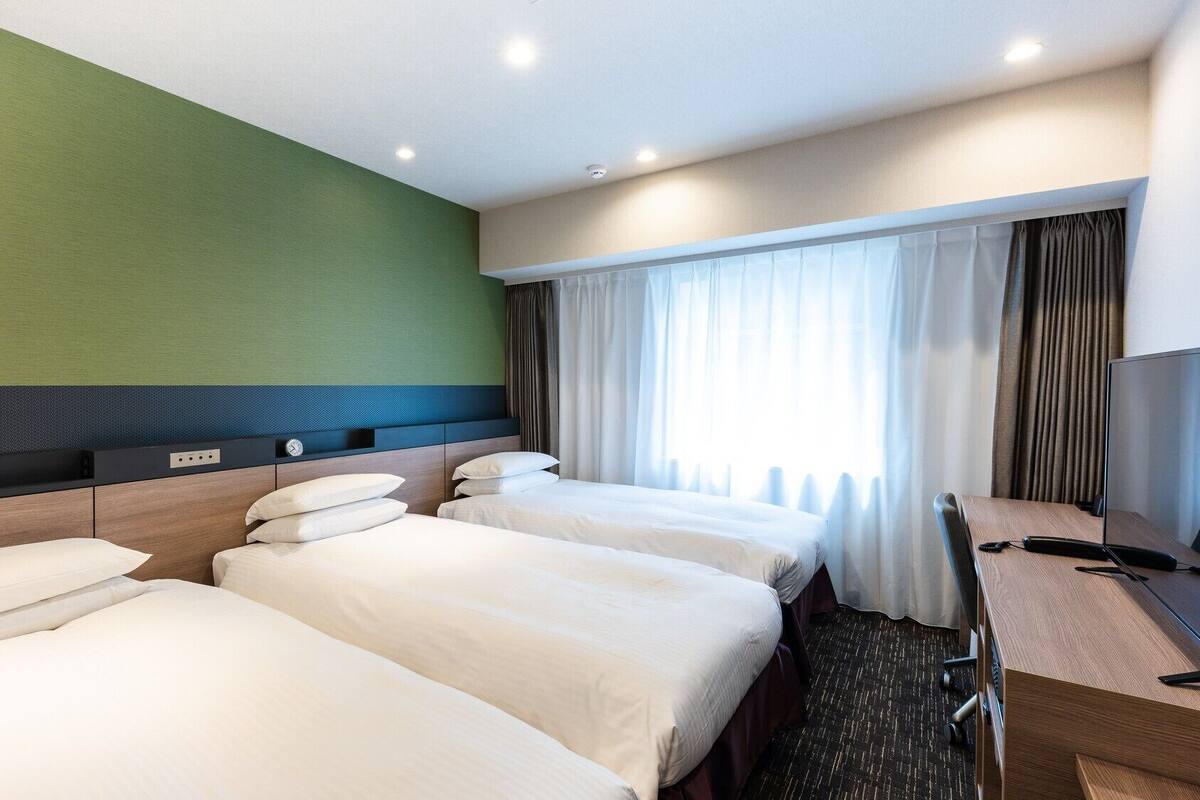 A hotel room with three beds, a desk, and a large window with curtains.