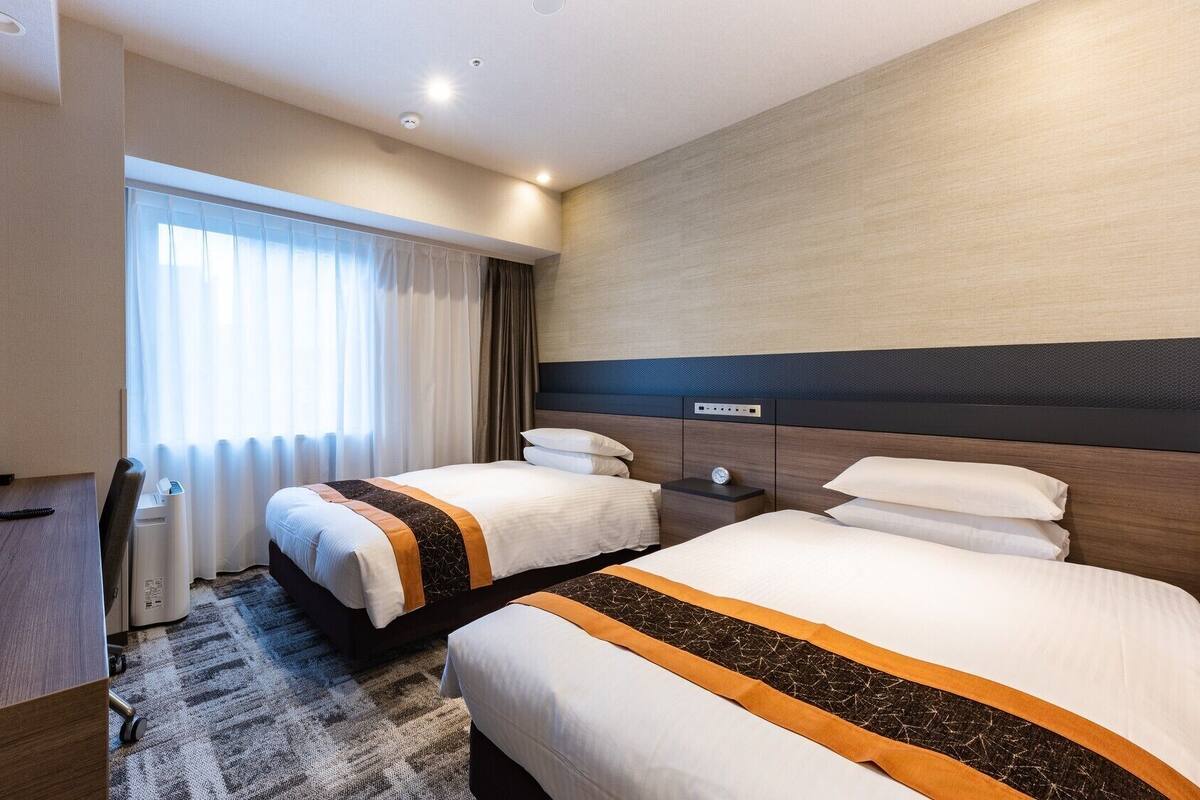 A hotel room with two beds, a desk, a TV, and a window with curtains.