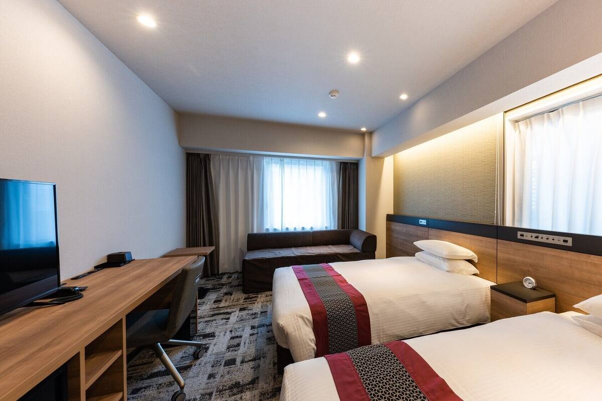 A hotel room with a bed, bedside table, TV, desk, and a window with curtains.