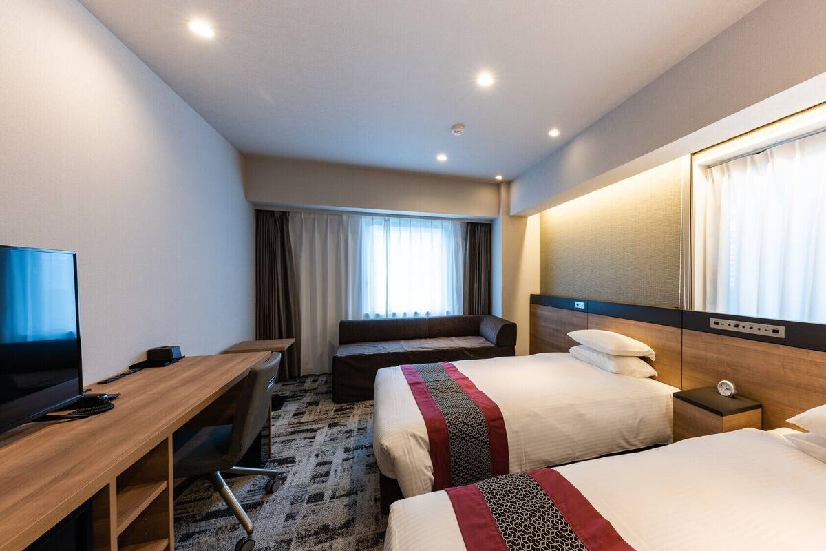 A hotel room with a bed, bedside table, TV, desk, and a window with curtains.
