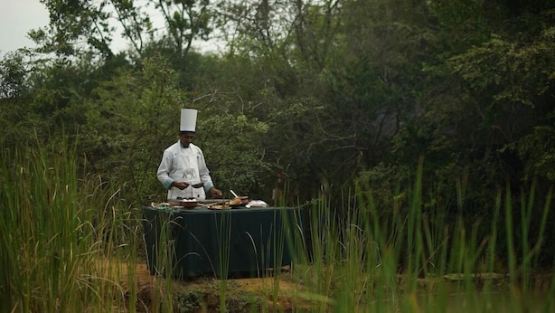 A chef in a white uniform with a tall hat, preparing food at a table set up in a natural, grassy area.