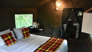 A tented accommodation room with a bed, a desk with a lamp, and a window with a view of greenery.