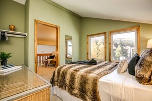 Un dormitorio con cama, escritorio y silla. Se ven ventanas con vistas a árboles y edificios.