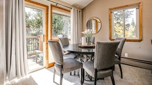 Un comedor con una mesa redonda, cuatro sillas y un florero con flores. Hay ventanas grandes con cortinas, y se ve un paisaje de árboles al exterior.